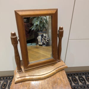 May include: A rectangular wooden swivel mirror featuring a light brown frame and a matching base. The mirror is supported by two vertical wooden posts on either side, allowing it to tilt. The wood has a visible grain and a smooth, polished finish.