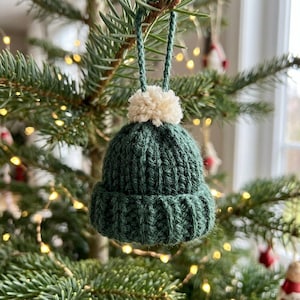 May include: A small, dark green knitted beanie ornament featuring a ribbed texture, a folded brim, and a fluffy cream-colored yarn pom-pom on top. A matching green yarn loop is attached to the crown for hanging purposes.