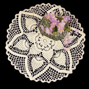 May include: A white crocheted doily with a delicate floral pattern. The doily is circular and has a scalloped edge. A bouquet of pink flowers is placed on top of the doily.