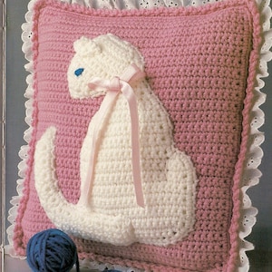 Könnte beinhalten: Ein rosa gehäkeltes Kissen mit einer weißen gehäkelten Katzenapplikation. Die Katze hat ein rosa Band um den Hals und blaue Augen. Das Kissen hat einen weißen Spitzenbesatz.