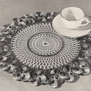 Può includere: Un centrino bianco all'uncinetto con un bordo a festoni e un disegno centrale. Il centrino è posto su un tavolo con un piattino bianco e una tazza da tè bianca.