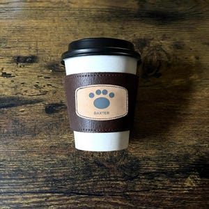 May include: A white disposable coffee cup with a black plastic lid and a dark brown textured leather sleeve. The sleeve features a centered light tan rectangular patch with a dark gray paw print icon and the text BAXTER printed below it.
