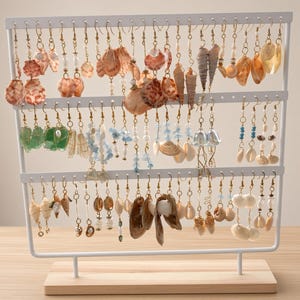 May include: A white metal earring display stand with a light wood base, holding numerous pairs of dangling earrings. The earrings feature various natural seashells, small white pearls, blue glass beads, and gold-toned wire wrapping on metal hooks.