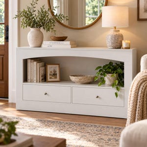 May include: A white wooden console table featuring an arched open shelf, two drawers with small round knobs, and a flat top surface. The table is styled with a speckled vase, a lamp, books titled The Kinfolk Home, a picture frame, and a bowl.