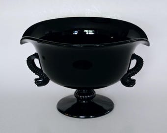 Puede incluir: Un cuenco de vidrio negro con dos asas en forma de caballitos de mar estilizados. El cuenco se asienta sobre una base de pedestal.