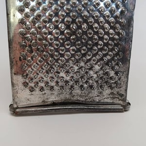 May include: A silver metal grater with a textured surface. The grater has a rectangular shape and a slightly curved top.