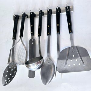 May include: A set of six silver kitchen utensils hanging on a black metal rack. The utensils include a potato masher, a spatula, a ladle, a spoon, a slotted spoon, and a slotted spatula.