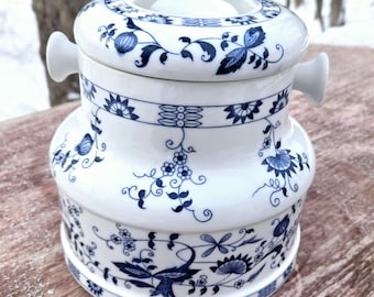 Puede incluir: Un recipiente de cerámica blanca con un patrón floral azul y blanco. El recipiente tiene una tapa con una pequeña manija a cada lado. El recipiente mide aproximadamente 15 cm de alto.