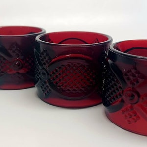 Puede incluir: Cuatro anillos de servilleta de vidrio rojo con un patrón decorativo. Los anillos están dispuestos en una fila sobre una superficie blanca.
