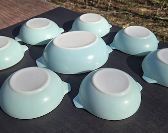 May include: A collection of eight light blue ceramic bowls with white interiors. Each bowl has a rounded shape with two small handles on opposite sides. The bowls are arranged on a dark surface, showcasing their vintage design.
