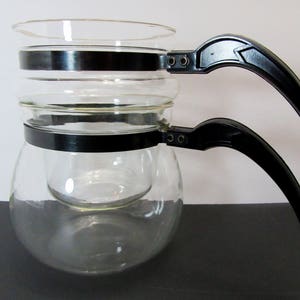 May include: A clear glass coffee pot with two black handles. The pot has a built-in filter and a glass carafe.