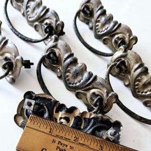 May include: Four antique drawer pulls with ornate, silver-painted metal designs and black metal handles. The pulls are arranged on a wooden surface with a ruler measuring 3 inches.