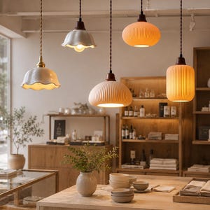 May include: Five hanging pendant lights with various shapes and textures, including white scalloped ceramic shades, ribbed orange glass, and a cylindrical ribbed glass shade, all suspended by dark twisted cords from a ceiling in a retail space.