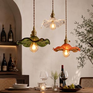 May include: Three pendant lights with ruffled glass shades in green, clear, and orange. Each lamp features a brass-toned socket and a brown twisted cord. The shades have a fluted, petal-like design with a glowing light bulb visible in each.