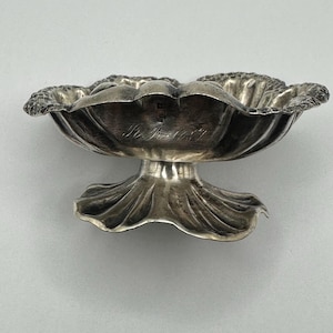 May include: A silver-toned metal bowl with a scalloped, shell-like design and a textured, ruffled rim. The bowl features a wide, flared base and an engraved cursive inscription on the interior side. The metallic surface shows reflective highlights.