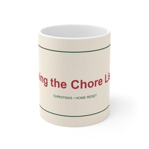 Könnte beinhalten: Ein cremefarbener Keramikbecher mit zwei dünnen dunkelgrünen horizontalen Linien oben und unten. Der rote Text lautet "ing the Chore Lis" und darunter steht in kleineren dunkelgrünen Großbuchstaben "CHRISTMAS • HOME RESET".