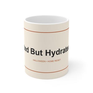 Könnte beinhalten: Ein cremefarbener Keramikbecher mit zwei dünnen horizontalen orangefarbenen Linien oben und unten. Der Text "ted But Hydrated" ist in Schwarz gedruckt, mit "HALLOWEEN • HOME RESET" in kleineren orangefarbenen Buchstaben unter dem Haupttext.