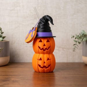 May include: A stack of two orange jack-o'-lanterns topped with a black witch hat featuring a purple band. A clear straw protrudes from the top, and a small orange pumpkin-shaped tag with a purple bow hangs from a silver chain on the side.