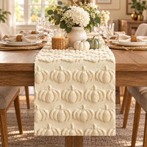 May include: A cream-colored table runner featuring a repeating embossed pumpkin pattern. The runner is made of a textured fabric and is draped over a wooden dining table, providing a neutral base for seasonal table decor and centerpieces.