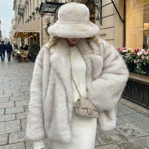 May include: A cream-coloured faux fur bucket hat and a matching cream faux fur coat worn over a cream-coloured knit dress. A small, quilted, cream-coloured crossbody bag with a gold-tone chain strap and a gold-tone logo emblem is worn over the shoulder.