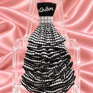 May include: A dress-shaped ornament featuring a black bodice with the word Queen in white script and a small crown icon. The waist is accented with a band of clear rhinestones. The skirt consists of multiple tiers of black and white gingham ruffles.