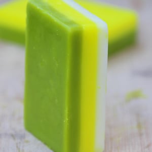 May include: A rectangular bar of soap featuring three distinct horizontal layers of color. The thickest layer is a solid light green, followed by a vibrant yellow middle section, and a thin white edge on the far side of the bar.