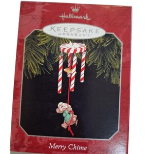 May include: A Hallmark Keepsake Ornament box featuring a Merry Chime wind chime ornament. The ornament has a red and white striped circular top with hanging candy cane style chimes and a small monkey figure in a green shirt hanging from a red cord.