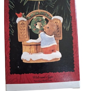 May include: A Hallmark Keepsake Ornament box featuring a brown bear wearing a blue jumper and orange trousers, standing at a wooden window frame with a green wreath, red berries, and snow. The box text reads Keepsake Ornament, Welcome Sign, Tender Touches.