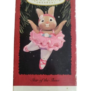 May include: A Hallmark Keepsake Ornament box featuring a brown rabbit ballerina wearing a pink tutu, a matching headband with a white flower, and pink ballet slippers. The box displays the text Keepsake Ornament and Star of the Show.
