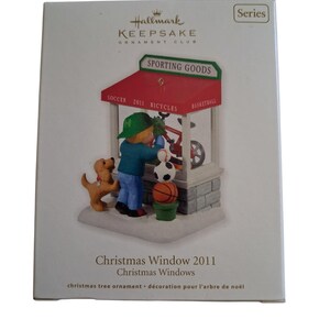 Könnte beinhalten: Ein Hallmark Keepsake Ornament Club Christmas Window 2011 Weihnachtsschmuck in seiner Verpackung. Das Ornament zeigt ein Geschäft mit der Aufschrift Sporting Goods, in dem ein Kind mit grünem Hut und blauem Oberteil ein Fahrrad betrachtet.