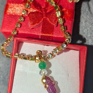 May include: A gold-tone chain necklace featuring clear rhinestones, a small green bead, a clear faceted bead, and a raw, deep pink-purple gemstone pendant. The necklace is displayed inside a small, square, bright red gift box with a ribbon.