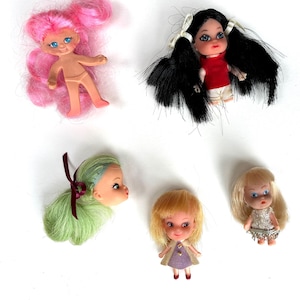 May include: A collection of five small vintage dolls with various hairstyles and outfits. One doll has pink hair, another has long black pigtails, and a third has green hair. The other two dolls have blonde hair and wear dresses.