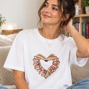 Peut inclure: Un t-shirt blanc à manches courtes avec une impression graphique de divers livres disposés en forme de cœur. Les livres sont représentés dans des tons marron, rose et crème, avec un livre ouvert au centre supérieur du motif en cœur.