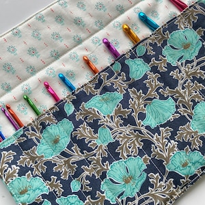 May include: A fabric crochet hook organizer case featuring a dark navy blue exterior with a teal and brown floral pattern and a cream-colored interior with a small teal and red geometric print, holding multiple colorful metal crochet hooks.