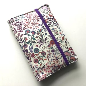 May include: A rectangular fabric notebook cover featuring a white background with a dense, colorful floral pattern in shades of purple, red, blue, and green. A single vertical purple elastic band secures the cover, which has stitched edges.