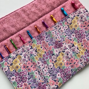 May include: A fabric crochet hook organizer case featuring a floral pattern in shades of pink, purple, yellow, and green. The interior contains multiple slots holding various metallic crochet hooks in vibrant colors like blue, orange, and pink.