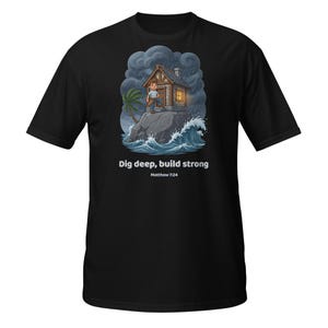 Op de afbeelding: Een zwart t-shirt met een afbeelding van een persoon die voor een huis op een rots staat tijdens een storm. De tekst "Dig deep, build strong" en "Matthew 7:24" staat in witte letters onder de illustratie van het huis en de golven.