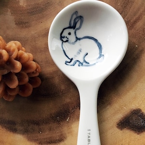 Puede incluir: Cuchara de cer&aacute;mica blanca con una ilustraci&oacute;n azul y blanca de un conejo en el cuenco. El mango de la cuchara es blanco y tiene el texto "1 TABLESPOON" impreso.