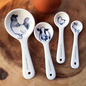 May include: Set of four white ceramic measuring spoons with animal illustrations. The spoons are labeled 1 tablespoon, 1 teaspoon, 1/2 teaspoon, and 1/4 teaspoon. The animals are a rooster, a cow, a duck, and a pig.