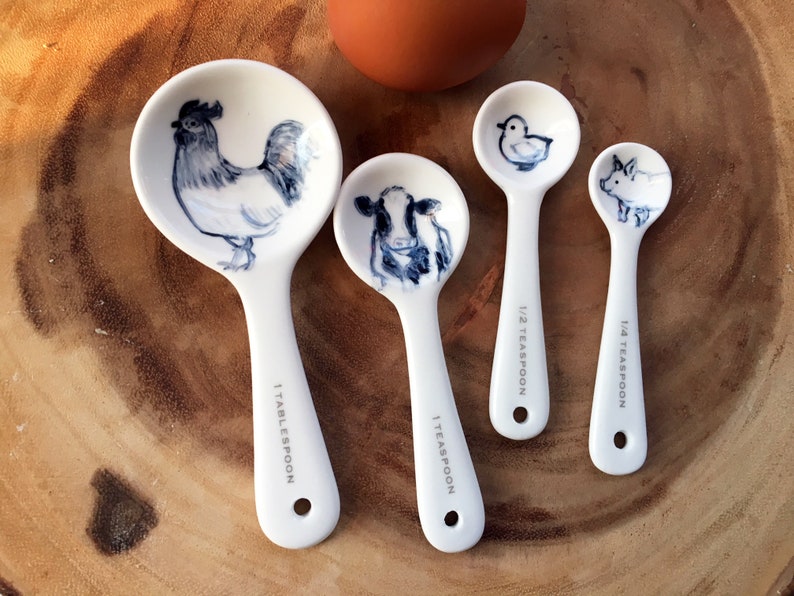 May include: Set of four white ceramic measuring spoons with animal illustrations. The spoons are labeled 1 tablespoon, 1 teaspoon, 1/2 teaspoon, and 1/4 teaspoon. The animals are a rooster, a cow, a duck, and a pig.