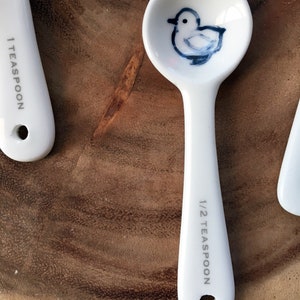 May include: White ceramic measuring spoon with a blue and white illustration of a bird. The spoon is marked "1/2 teaspoon".