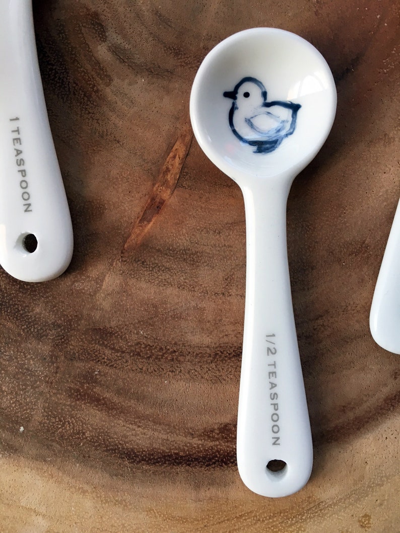 May include: White ceramic measuring spoon with a blue and white illustration of a bird. The spoon is marked "1/2 teaspoon".