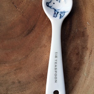 May include: White ceramic teaspoon with a blue and white illustration of a pig on the bowl. The handle is marked "1/4 Teaspoon".