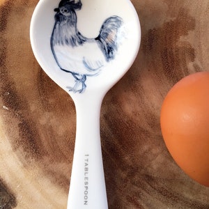 May include: White ceramic measuring spoon with a blue and white rooster illustration. The spoon is marked "1 Tablespoon".