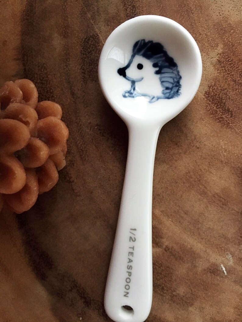 Puede incluir: Cuchara de t&eacute; de cer&aacute;mica blanca con un dise&ntilde;o de erizo azul y blanco. La cuchara est&aacute; marcada con "1/2 cucharadita".