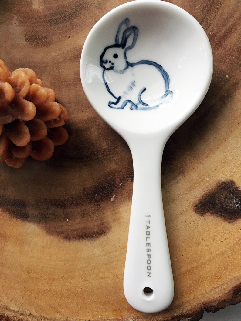 Puede incluir: Cuchara de cer&aacute;mica blanca con una ilustraci&oacute;n azul y blanca de un conejo en el cuenco. El mango de la cuchara es blanco y tiene el texto "1 TABLESPOON" impreso.