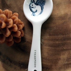 Puede incluir: Una cucharadita de cer&aacute;mica blanca con una ilustraci&oacute;n de ardilla azul y blanca en el cuenco. El mango est&aacute; marcado con "1 TEASPOON".