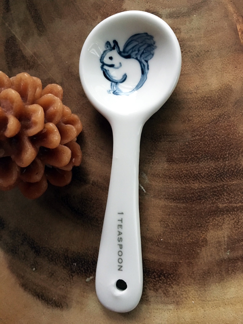 Puede incluir: Una cucharadita de cer&aacute;mica blanca con una ilustraci&oacute;n de ardilla azul y blanca en el cuenco. El mango est&aacute; marcado con "1 TEASPOON".