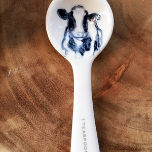 May include: White ceramic teaspoon with a black and white cow illustration on the bowl. The handle is marked "1 TEASPOON".