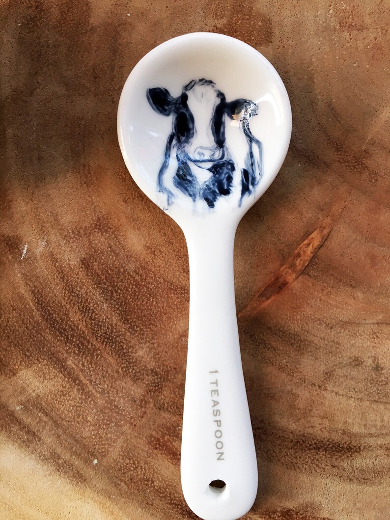May include: White ceramic teaspoon with a black and white cow illustration on the bowl. The handle is marked "1 TEASPOON".